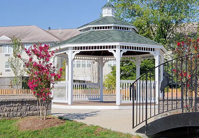A white gazebo sitting in a peaceful setting with a red flowering tree in the foreground.